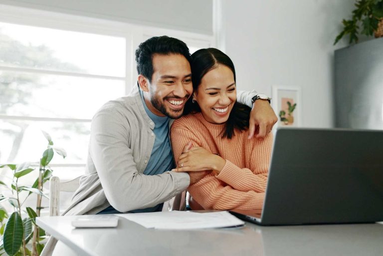 Couple in kitchen smiling at laptop tracking expenses for budget goals.