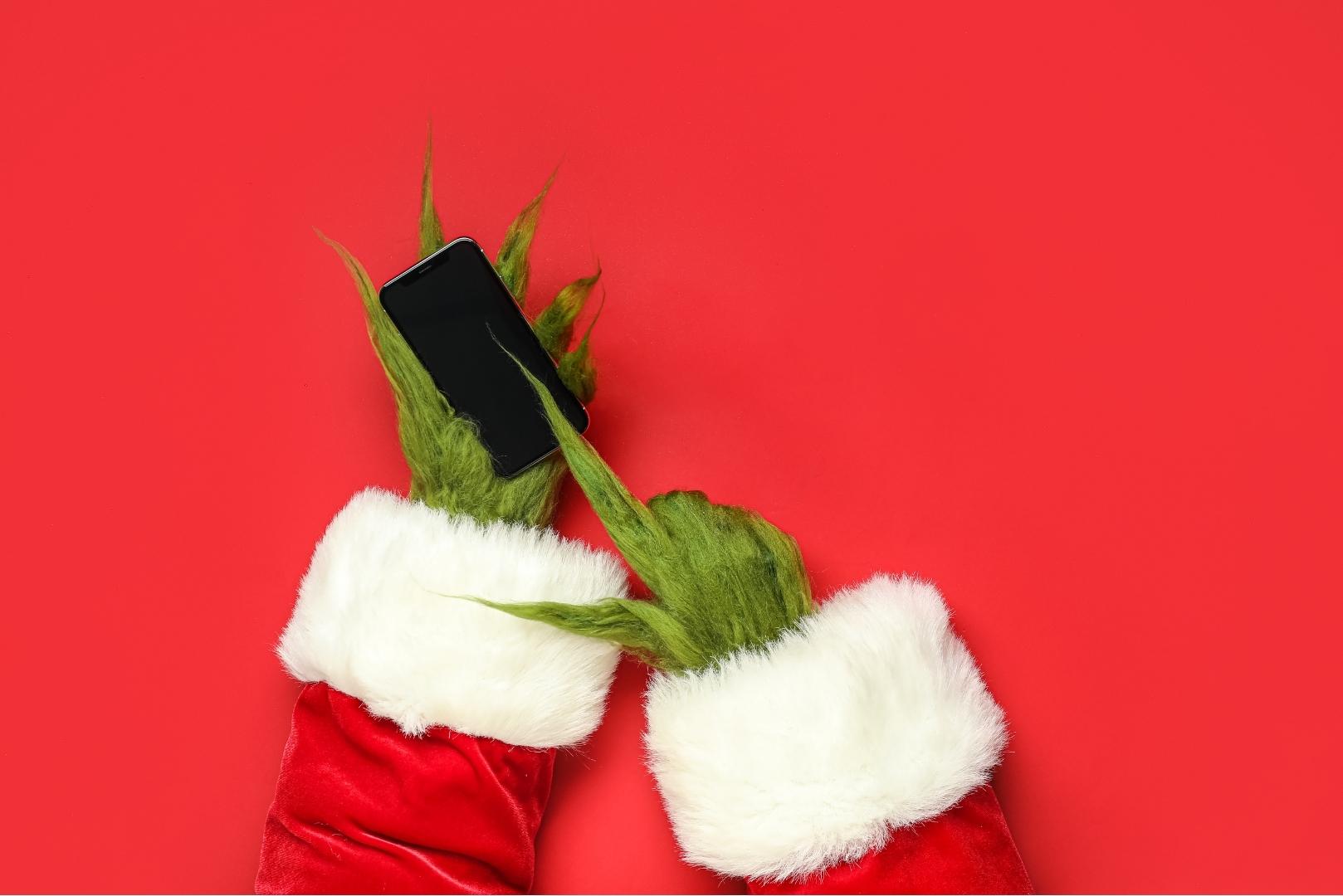 Two green, furry hands wearing red Santa cuffs hold a black smartphone against a red background.
