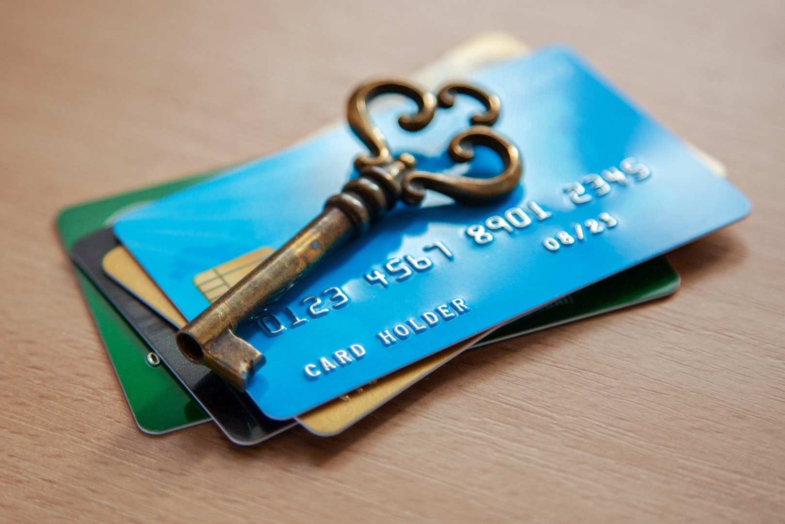 A vintage brass key rests on a stack of credit cards, with the top card being blue and displaying embossed numbers and printed numbers and text.