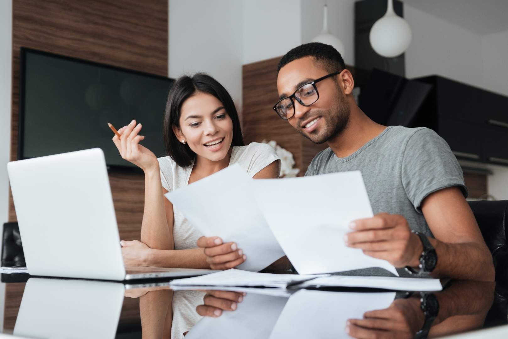 A young couple sitting at a table looking at papers and a laptop is to the left of the woman.