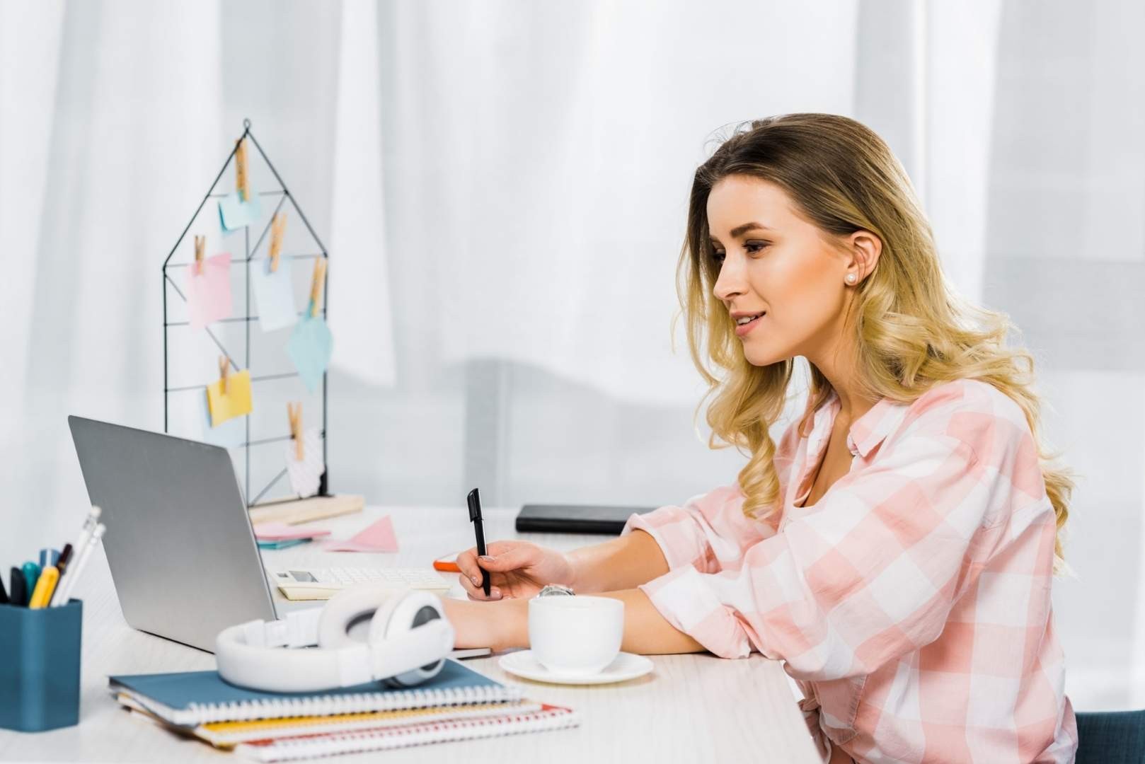 A woman is sitting at a desk and writing in a notebook while looking at a laptop.