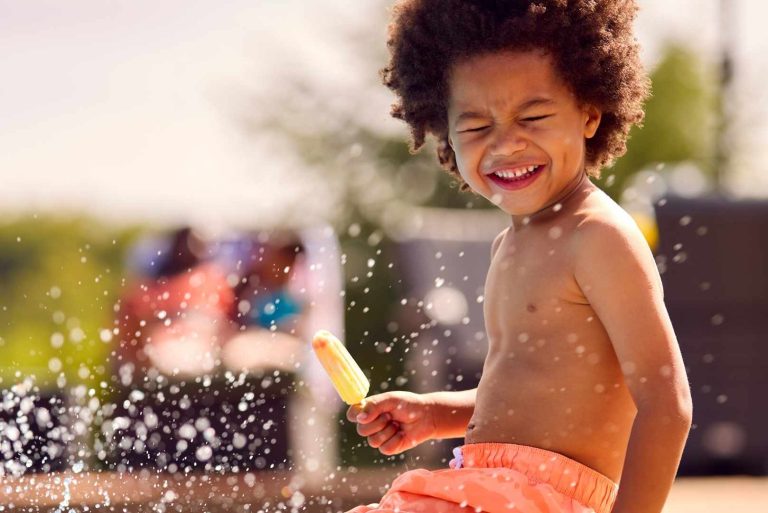 A child with curly hair laughs while holding a popsicle and being sprayed with water.