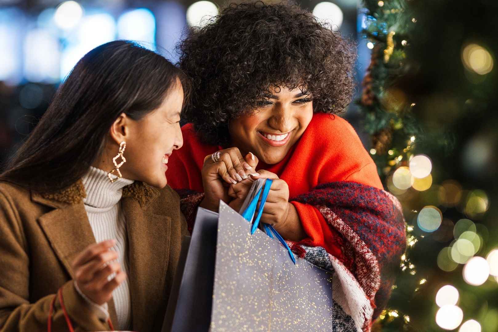 Two women smiling and leaning in while holding gift bags; with soft holiday lights in the background.